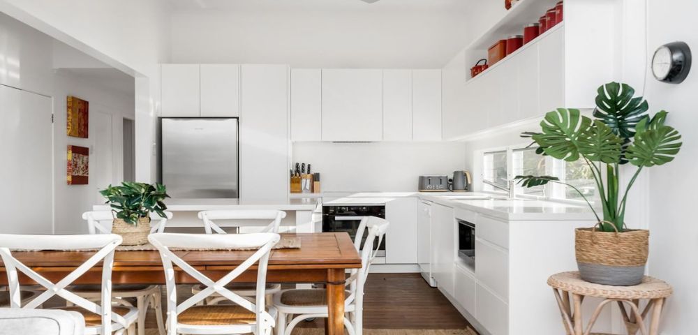 Bright, modern kitchen with white cabinets, stainless steel fridge, wooden dining table, white chairs, green plants, and red decor accents on shelves.v