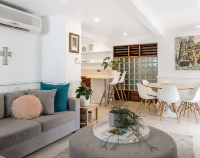 Bright, modern living room with gray sofa, pink and teal cushions, round coffee table, dining area with white chairs, and vibrant abstract art. Cozy ambiance.