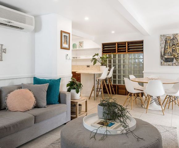 Bright, modern living room with gray sofa, pink and teal cushions, round coffee table, dining area with white chairs, and vibrant abstract art. Cozy ambiance.
