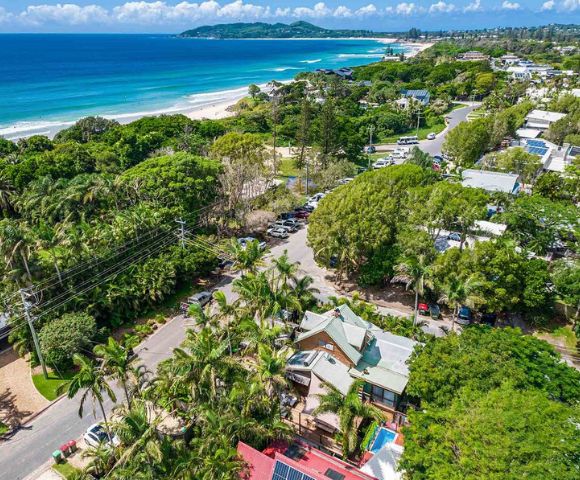 Aerial view of a coastal town with lush greenery, palm trees, and houses. The ocean nearby has blue waves, and the sky is clear, conveying a serene atmosphere.