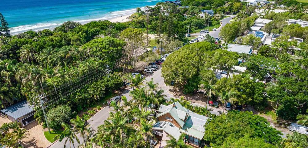Aerial view of a coastal town with lush greenery, palm trees, and houses. The ocean nearby has blue waves, and the sky is clear, conveying a serene atmosphere.