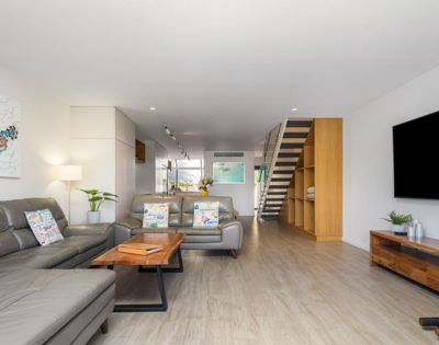 Modern living room with a gray sectional sofa, wooden coffee table, and flat-screen TV. Bright, open space with hardwood floors and staircase.