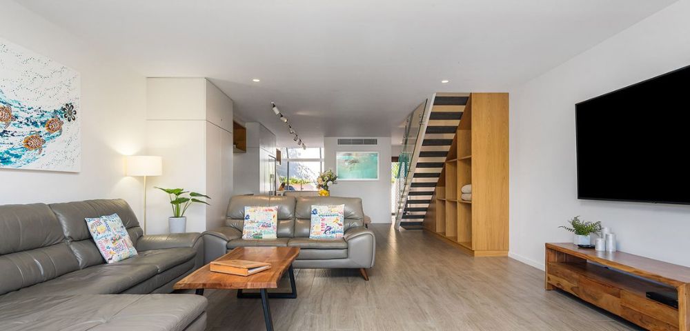 Modern living room with a gray sectional sofa, wooden coffee table, and flat-screen TV. Bright, open space with hardwood floors and staircase.