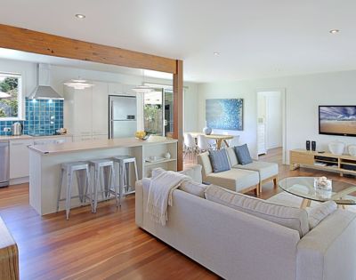 Modern open-plan living area with light wood floors, a cozy white sofa, blue accents, a kitchen island with stools, and a dining table. Airy and inviting atmosphere.