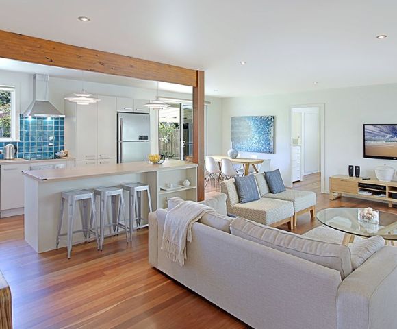 Modern open-plan living area with light wood floors, a cozy white sofa, blue accents, a kitchen island with stools, and a dining table. Airy and inviting atmosphere.