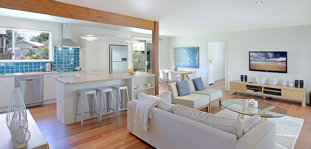 Modern open-plan living area with light wood floors, a cozy white sofa, blue accents, a kitchen island with stools, and a dining table. Airy and inviting atmosphere.
