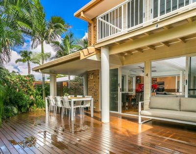 A sunlit patio featuring a polished wooden deck, outdoor dining set, and cushioned seating. Surrounded by lush tropical plants, the scene is serene and inviting.