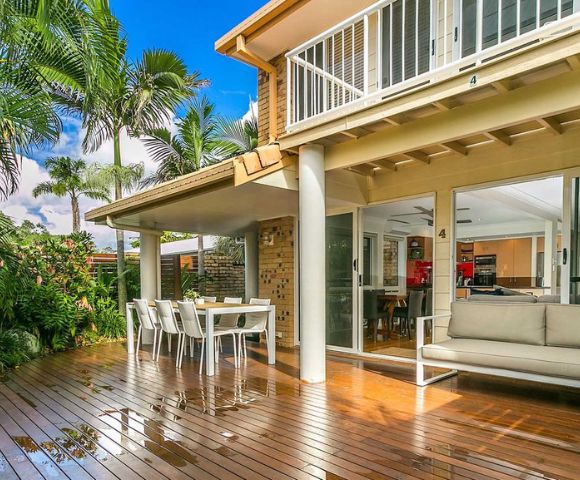 A sunlit patio featuring a polished wooden deck, outdoor dining set, and cushioned seating. Surrounded by lush tropical plants, the scene is serene and inviting.