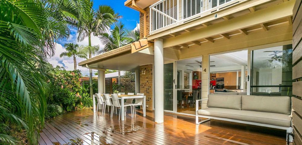 A sunlit patio featuring a polished wooden deck, outdoor dining set, and cushioned seating. Surrounded by lush tropical plants, the scene is serene and inviting.