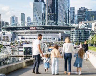 A family of five walks on a riverside promenade toward a stadium in a cityscape. The scene is sunny, with modern skyscrapers in the background.