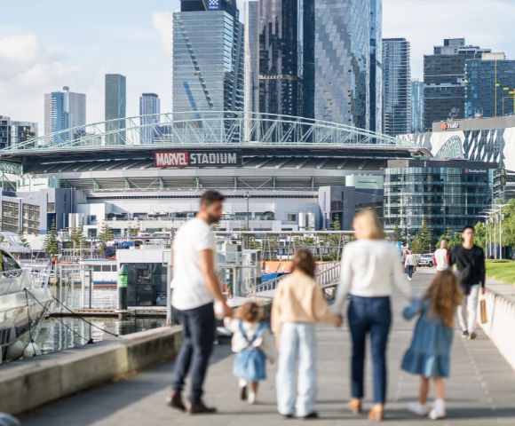 A family of five walks on a riverside promenade toward a stadium in a cityscape. The scene is sunny, with modern skyscrapers in the background.