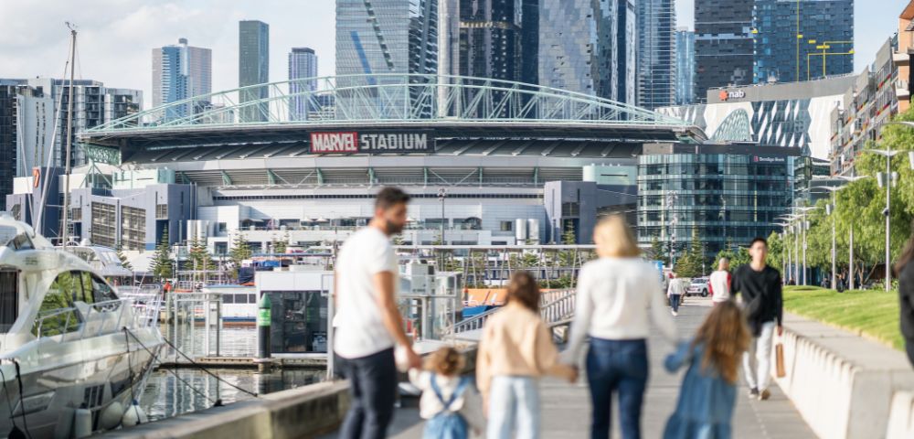 A family of five walks on a riverside promenade toward a stadium in a cityscape. The scene is sunny, with modern skyscrapers in the background.