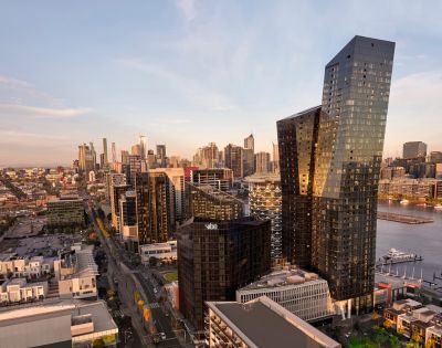 Aerial view of a modern cityscape at sunset, featuring sleek skyscrapers with reflective glass, a river, and warm light casting long shadows.