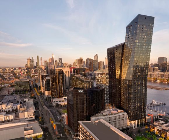 Aerial view of a modern cityscape at sunset, featuring sleek skyscrapers with reflective glass, a river, and warm light casting long shadows.