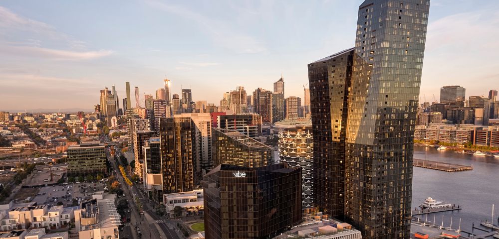 Aerial view of a modern cityscape at sunset, featuring sleek skyscrapers with reflective glass, a river, and warm light casting long shadows.