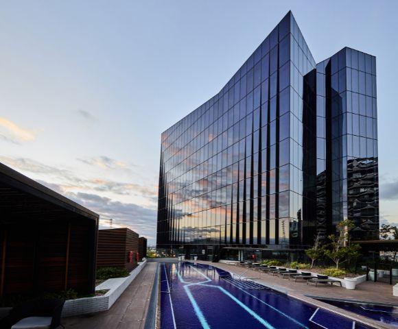 Modern glass building reflecting the sunset sky stands beside a sleek rooftop pool with lounge chairs, creating a serene and luxurious atmosphere.