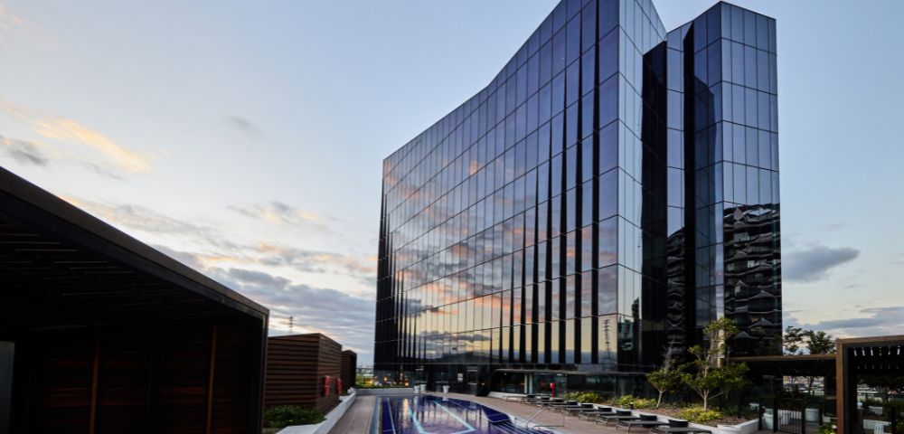 Modern glass building reflecting the sunset sky stands beside a sleek rooftop pool with lounge chairs, creating a serene and luxurious atmosphere.