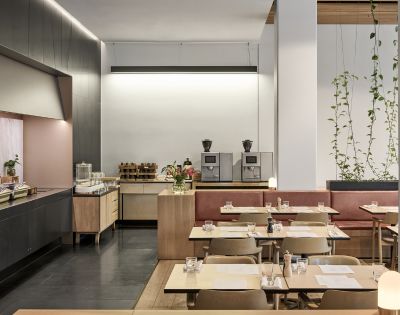 Modern café interior with light wooden tables and chairs, red banquette seating, and greenery. Coffee machines and pastries on a wooden counter create a cozy ambiance.