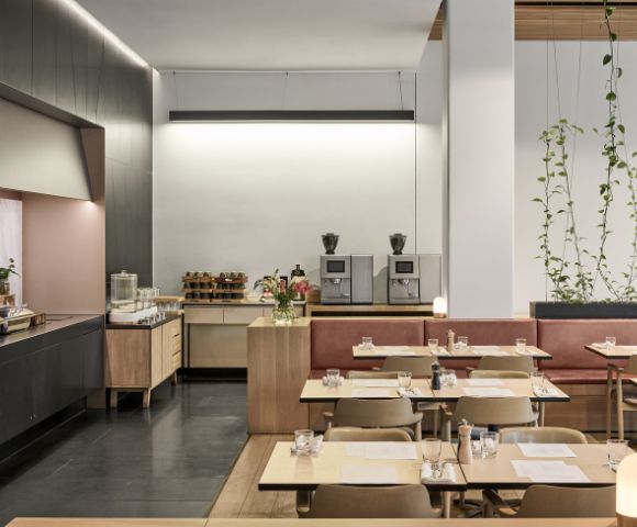 Modern café interior with light wooden tables and chairs, red banquette seating, and greenery. Coffee machines and pastries on a wooden counter create a cozy ambiance.