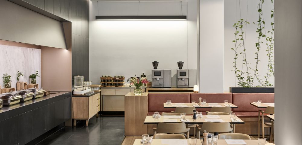 Modern café interior with light wooden tables and chairs, red banquette seating, and greenery. Coffee machines and pastries on a wooden counter create a cozy ambiance.