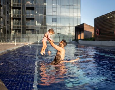 A man joyfully lifts a child in a rooftop pool, with a woman nearby. Modern glass buildings in the background under a clear blue sky.