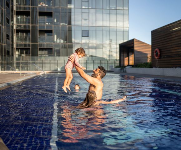 A man joyfully lifts a child in a rooftop pool, with a woman nearby. Modern glass buildings in the background under a clear blue sky.