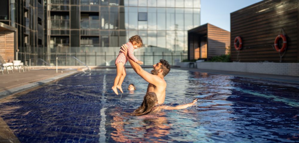 A man joyfully lifts a child in a rooftop pool, with a woman nearby. Modern glass buildings in the background under a clear blue sky.