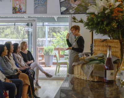 A woman in an apron gestures while speaking to an attentive seated group. Nearby is a basket with flowers and a wine bottle. The setting is cozy and inviting.