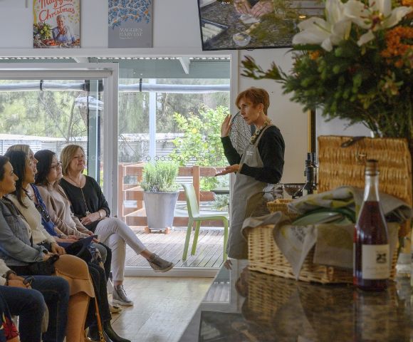 A woman in an apron gestures while speaking to an attentive seated group. Nearby is a basket with flowers and a wine bottle. The setting is cozy and inviting.