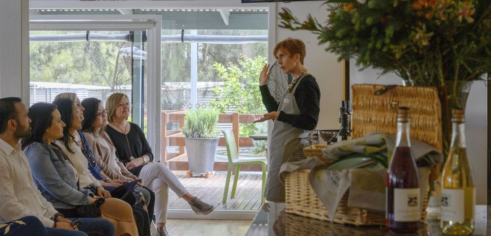 A woman in an apron gestures while speaking to an attentive seated group. Nearby is a basket with flowers and a wine bottle. The setting is cozy and inviting.