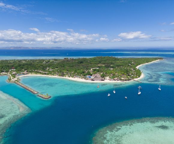 Aerial view of a lush tropical island surrounded by turquoise waters, white sandy beaches, and several anchored sailboats under a clear blue sky.