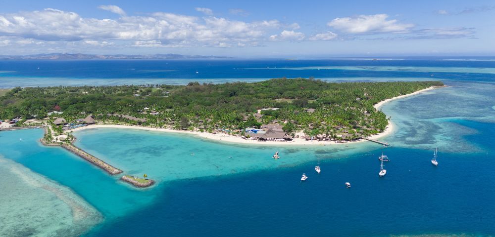 Aerial view of a lush tropical island surrounded by turquoise waters, white sandy beaches, and several anchored sailboats under a clear blue sky.