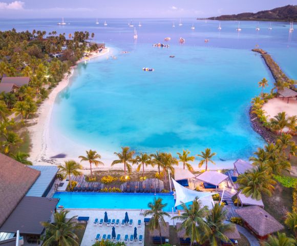 Aerial view of a tropical beach with turquoise water, palm-lined shore, and sailboats. The scene is serene, evoking a tranquil, vacation vibe.