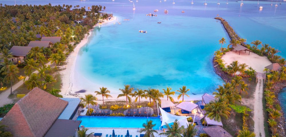 Aerial view of a tropical beach with turquoise water, palm-lined shore, and sailboats. The scene is serene, evoking a tranquil, vacation vibe.