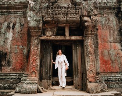 A person in white clothing stands at an intricately carved, ancient temple doorway. The weathered stone facade is rich with earthy tones and textures.