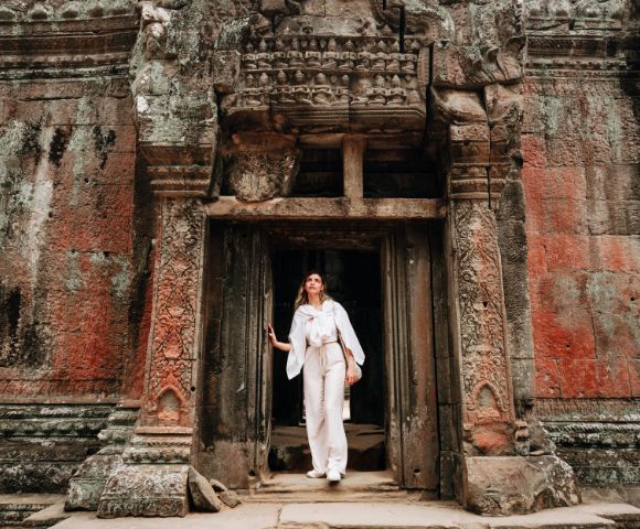 A person in white clothing stands at an intricately carved, ancient temple doorway. The weathered stone facade is rich with earthy tones and textures.