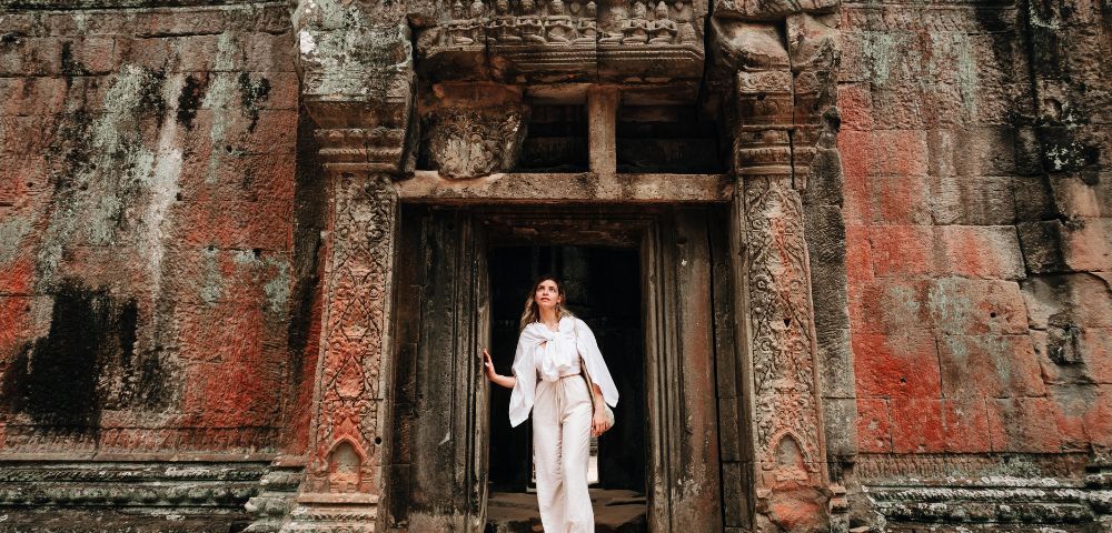 A person in white clothing stands at an intricately carved, ancient temple doorway. The weathered stone facade is rich with earthy tones and textures.