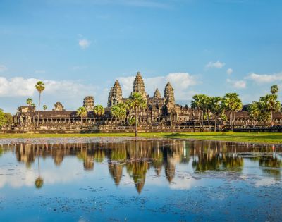 Ancient temple with tall spires reflected in a calm pond under a clear blue sky. Surrounded by lush greenery and palm trees, creating a serene atmosphere.
