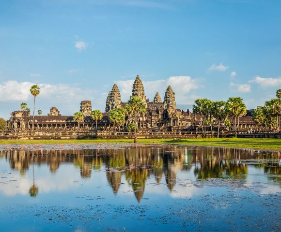 Ancient temple with tall spires reflected in a calm pond under a clear blue sky. Surrounded by lush greenery and palm trees, creating a serene atmosphere.