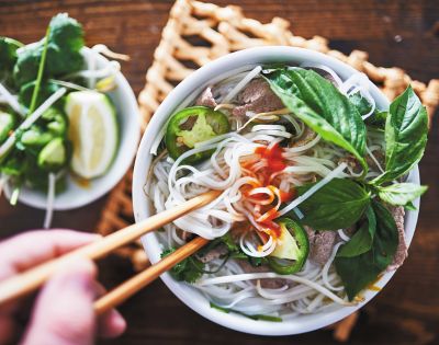 A steaming bowl of Vietnamese pho with noodles, beef slices, jalapeños, and fresh basil, garnished with chili sauce. A hand uses chopsticks to lift noodles.