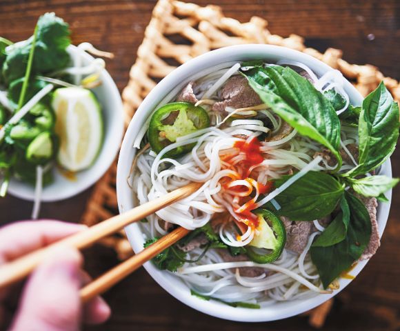 A steaming bowl of Vietnamese pho with noodles, beef slices, jalapeños, and fresh basil, garnished with chili sauce. A hand uses chopsticks to lift noodles.