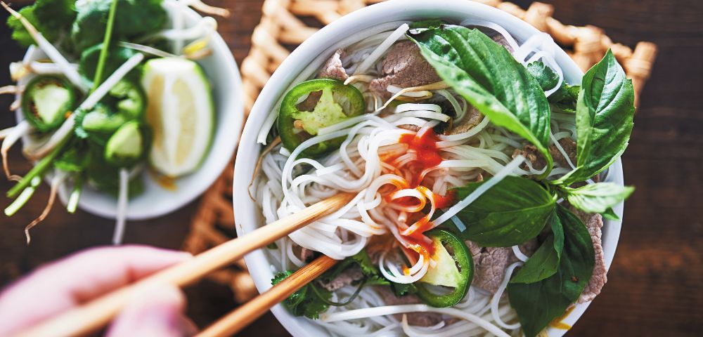 A steaming bowl of Vietnamese pho with noodles, beef slices, jalapeños, and fresh basil, garnished with chili sauce. A hand uses chopsticks to lift noodles.