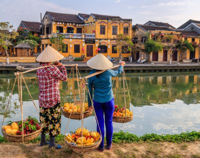 Two people in conical hats stand by a tranquil river, carrying baskets of vibrant fruits. Historic yellow buildings line the opposite bank, reflecting cultural charm.