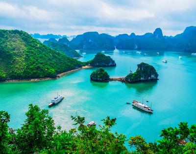 Serene view of Halong Bay with turquoise water, lush green islands, and boats. Overcast sky adds a calming, picturesque atmosphere to the landscape.
