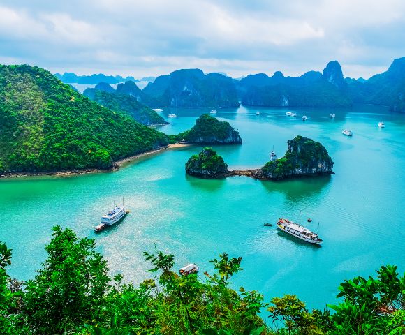 Serene view of Halong Bay with turquoise water, lush green islands, and boats. Overcast sky adds a calming, picturesque atmosphere to the landscape.