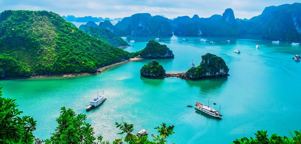 Serene view of Halong Bay with turquoise water, lush green islands, and boats. Overcast sky adds a calming, picturesque atmosphere to the landscape.