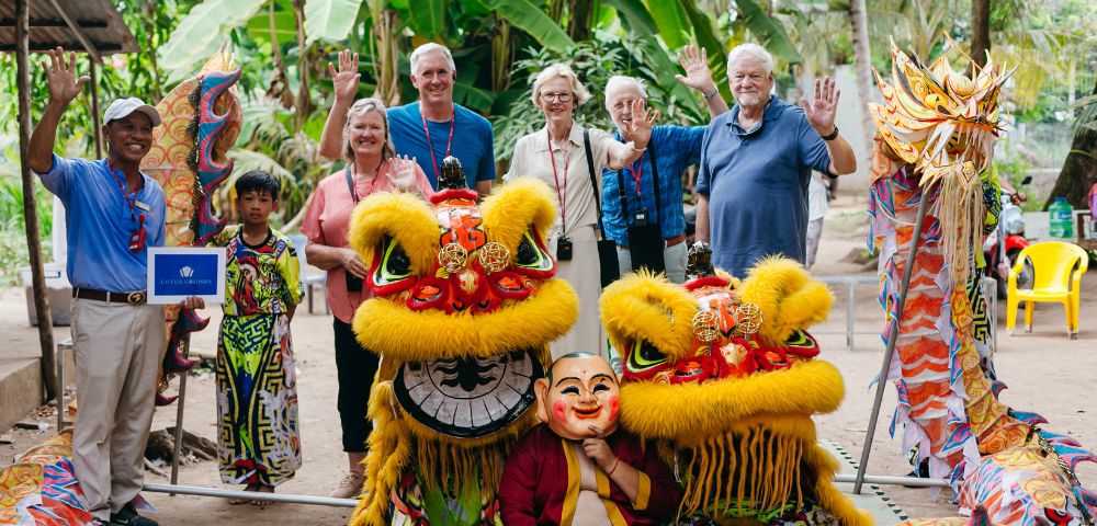 A group of smiling people and two lion dancers in colorful costumes pose outdoors with lush greenery. They wave enthusiastically, creating a joyful atmosphere.