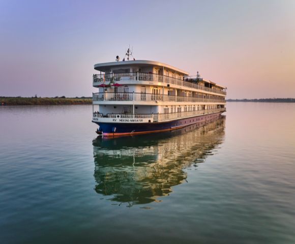 A large white cruise ship glides on calm waters at sunset, reflecting in the serene river. The sky is pastel, evoking a peaceful and tranquil mood.