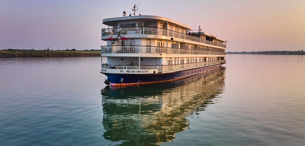 A large white cruise ship glides on calm waters at sunset, reflecting in the serene river. The sky is pastel, evoking a peaceful and tranquil mood.