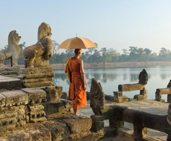 A monk in orange robes stands on ancient stone steps by a tranquil lake, holding a beige umbrella. Nearby are old lion statues. The mood is serene and reflective.